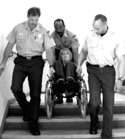 Black and white photograph showing the correct technique for assisting the evacuation of a person who uses a wheelchair. U.S. Fire Administration 'Orientation Manual for First Responders on the Evacuation of People with Disabilities'. FA-235/August 2002.