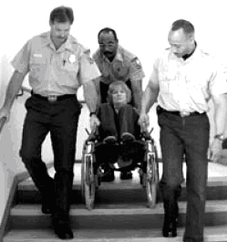 Black and white photograph (US FEMA 2002) showing the correct way to assist the fire evacuation of a wheelchair user in an evacuation staircase ... one person at each side, with another person behind.