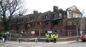 Colour photograph showing the cordoned-off scene in the aftermath of the fires at a Terrace of Housing in Terenure, Dublin City. In the foreground, Gardaí are keeping a watchful eye. Photograph taken by CJ Walsh. 2011-04-04. Click to enlarge.