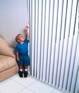 Colour photograph showing a small child reaching for the control wand of a Vertical Window Blind in familiar surroundings, for example, at home. This is just one of a range of Child Safe Window Covering Solutions.